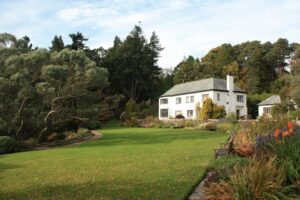 White colonial-style home surrounded by mature trees, manicured lawn, and flower gardens on a hillside estate.